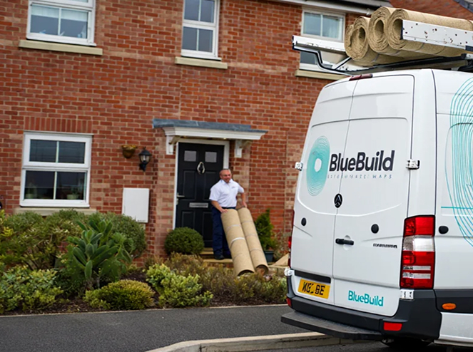 A rear close-up view of a white branded BlueBuild service van parked on the street with insulation rolls stacked on its roof rack, while a team member in a polo shirt carries rolls of insulation toward the front door of a red brick residential house.