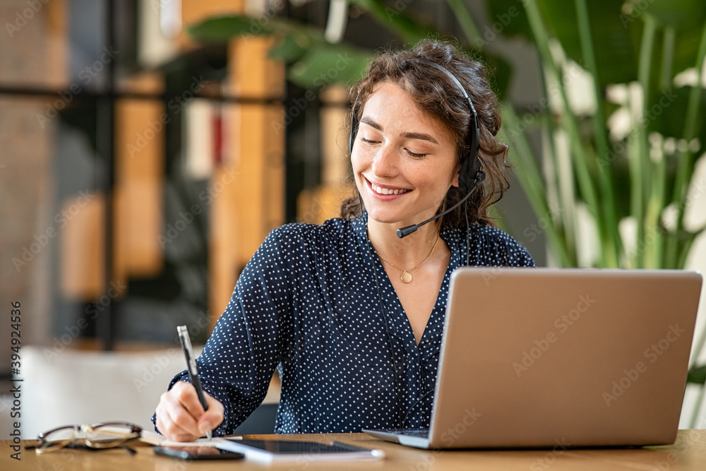 A smiling woman wearing a headset takes notes with a pen while looking toward her open laptop at a wooden desk in a bright office environment.