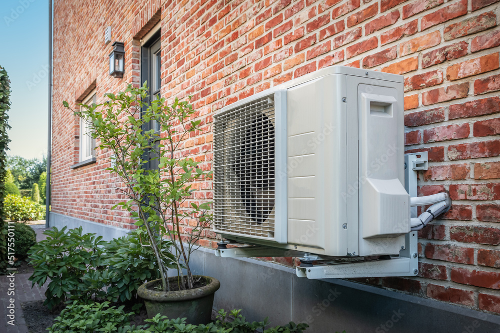 An external residential air-to-water heat pump compressor unit mounted on heavy-duty wall brackets against a red brick house facade, surrounded by neat garden bushes under warm sunlight.