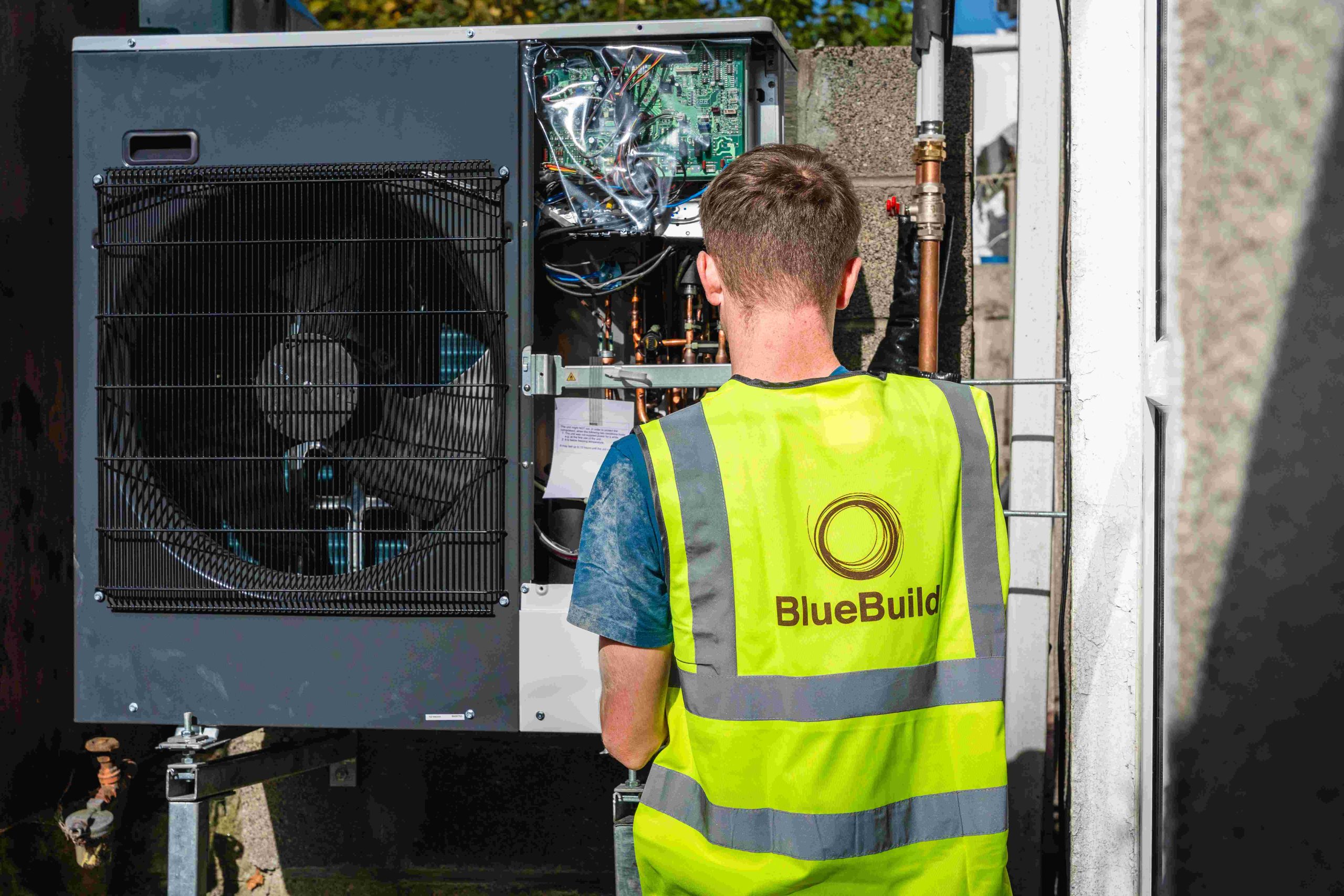 A rear view of a BlueBuild technician wearing a high-visibility vest as he works on the internal wiring and components of an outdoor residential air-to-water heat pump system during installation.