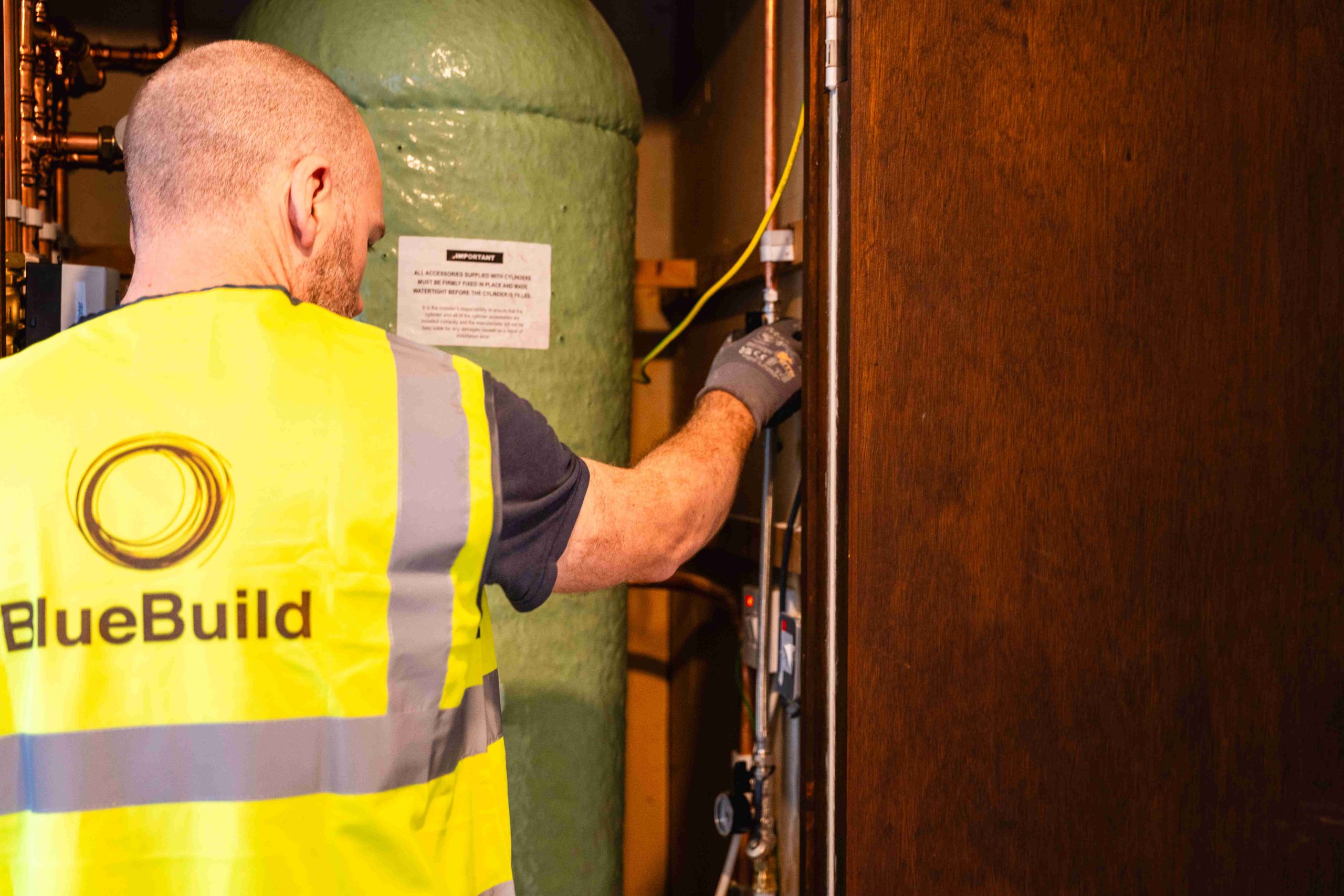 A rear view of a BlueBuild technician wearing a high-visibility vest as he reaches inside a hot press cupboard to adjust plumbing valves and copper pipework next to a green insulated hot water cylinder.